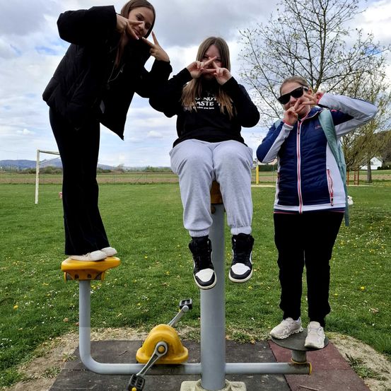 Drei Mädchen posieren für ein Foto in einem Park, eine von ihnen sitzt auf einem Wippgerät, während die anderen stehen, alle lächeln und Friedenszeichen machen.