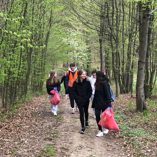 Eine Gruppe von Menschen geht einen schlammigen Pfad in einem Wald entlang. Sie tragen Jacken und tragen rote Taschen.