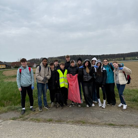 Eine Gruppe von Schülern, die Handschuhe tragen und Plastiktüten halten, steht auf einem Feld. Der Hintergrund zeigt einen bewölkten Himmel, ein Grasfeld und ein gelbes Haus.