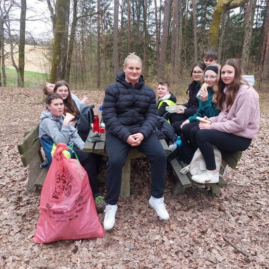 Eine Gruppe junger Menschen sitzt auf einer Holzbank im Wald, einer isst Essen und einer hat einen rosa Beutel auf dem Boden.