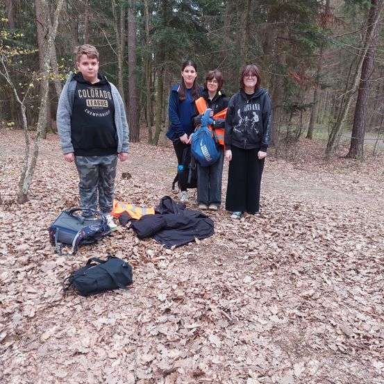 Vier Menschen stehen in einem Wald, zwei davon sind Frauen und zwei sind Jungen. Alle tragen Jacken und halten einen Rucksack. Es liegen Kleidungsstücke und Taschen auf dem Boden.