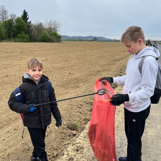 Zwei Jungen halten Plastiktüten auf einem Feldweg mit einem Feld im Hintergrund. Ein Junge hält einen Stock mit Plastiktüten am Ende.