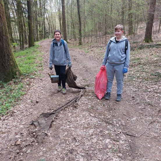 Zwei Jungen gehen auf einem Waldweg. Einer hält einen Eimer, der andere einen roten Plastikbeutel.