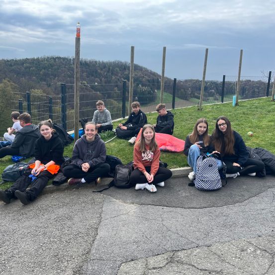 Eine Gruppe junger Menschen sitzt auf dem Rasen vor einem Zaun mit Blick auf einen Wald und Berge.