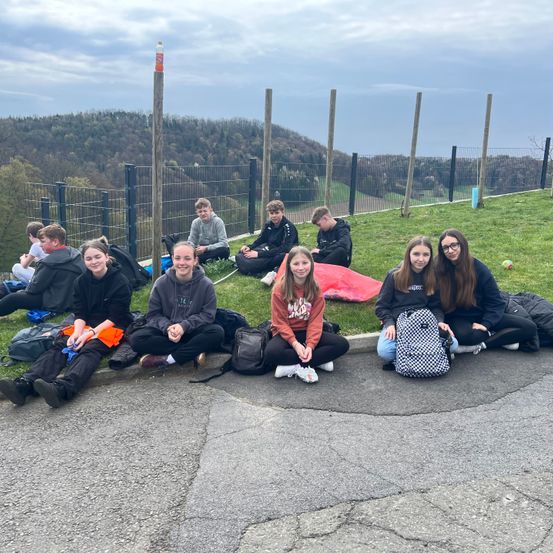 Sieben junge Menschen sitzen am Straßenrand, in der Nähe eines Rasens mit Zaun und Blick auf die Berge.