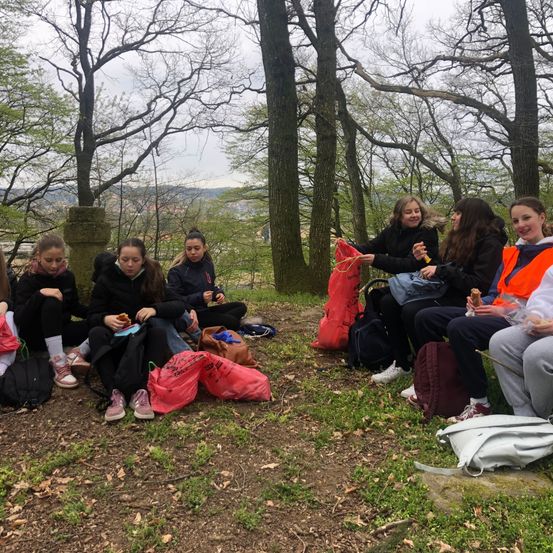 Eine Gruppe junger Mädchen sitzt auf einem Rasen in einem Wald. Sie essen und plaudern. Jedes Mädchen hat einen Rucksack, und einige tragen zusätzliche Taschen. Bäume umgeben sie, und in der Ferne ist eine Bergkette zu sehen.