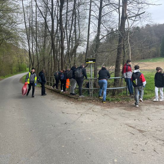 Eine Gruppe junger Leute steht am Straßenrand, einige tragen Rucksäcke und andere halten Plastiktüten. Sie schauen auf etwas im Feld auf der anderen Seite einer Metallbarriere.