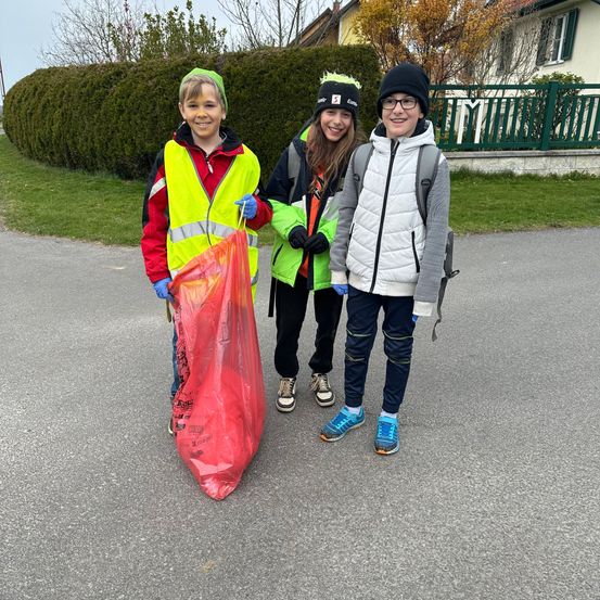 Drei Kinder stehen lächelnd auf der Straße. Eines von ihnen hält einen roten Plastikbeutel. Sie tragen Handschuhe und Sneakers. Der Junge links trägt eine gelbe Jacke, während die anderen beiden grüne Jacken tragen. Dahinter steht ein Busch und ein Haus mit einem Zaun.
