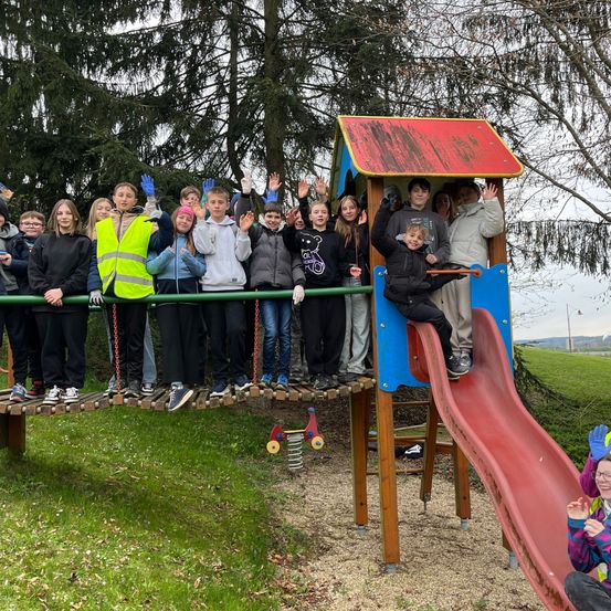 Eine Gruppe von Kindern in Winterkleidung steht auf einem Spielplatz mit einer Rutsche und einer kleinen Hütte.