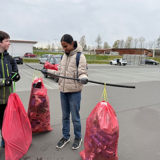 Zwei junge Leute tragen Plastiktüten mit Plastikflaschen in einem Außenbereich, wahrscheinlich einem Parkplatz. Die Person links trägt einen schwarzen Mantel mit grünem Reißverschluss, während die andere einen beigen Mantel und Jeans trägt.