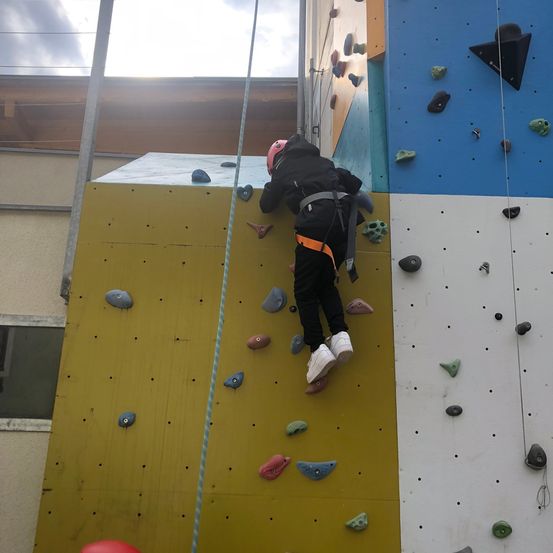 Ein Kind klettert an einer Indoor-Kletterwand, trägt einen Helm und Turnschuhe und hat einen Sicherheitsgurt am Seil befestigt.