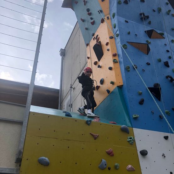 Eine Person klettert an einer Kletterwand mit bunten Griffen und Tritthilfen und trägt einen Helm und Turnschuhe.