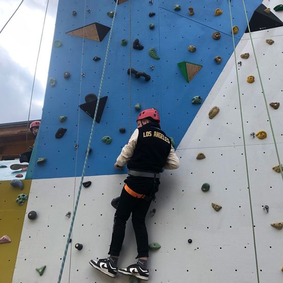 Eine Person mit rotem Helm und schwarzem Weste mit Los Angeles Aufschrift klettert an einer Kletterwand mit verschiedenfarbigen Steinen.