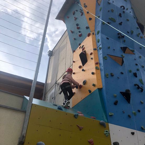 Ein junges Mädchen mit rotem Helm klettert an einer mehrfarbigen Kletterwand mit einem Netzzaun im Hintergrund.