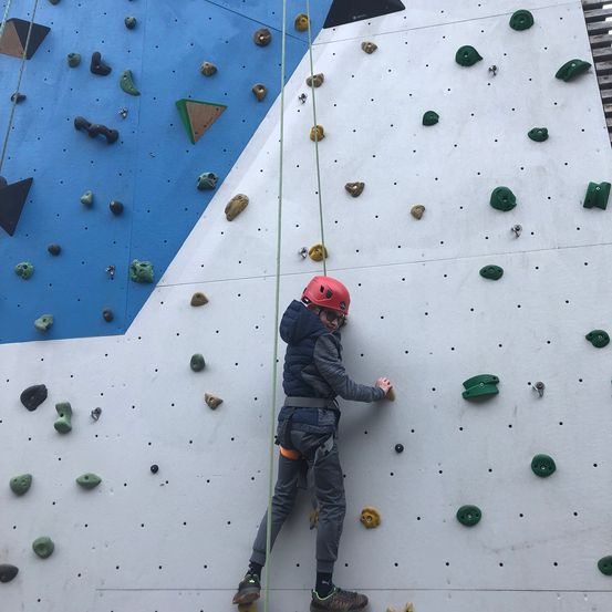 Eine Person mit rotem Helm und Gurt klettert an einer weißen Kletterwand mit grünen und gelben Griffen hoch.
