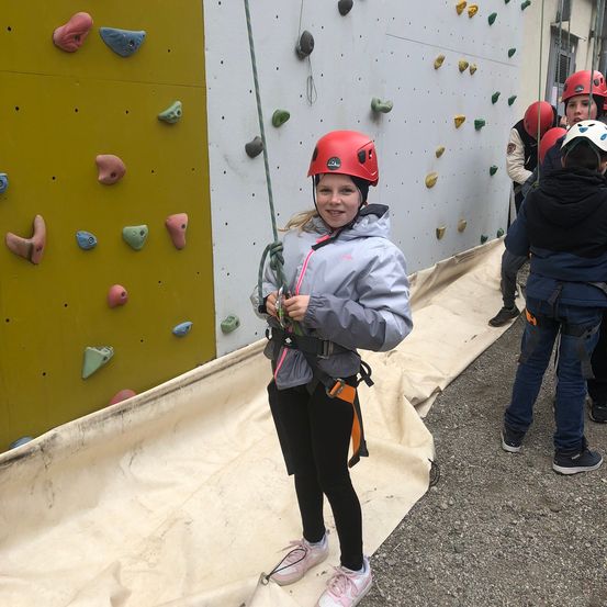 Ein junges Mädchen mit rotem Helm und Gurt steht neben einer Kletterwand mit farbigen Griffen.