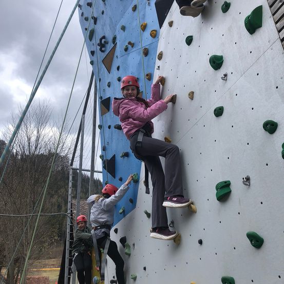 Zwei Frauen in roten Helmen klettern an einer Indoor-Kletterwand, eine Frau lächelt und schaut nach unten, während die andere ihr Bein hält.