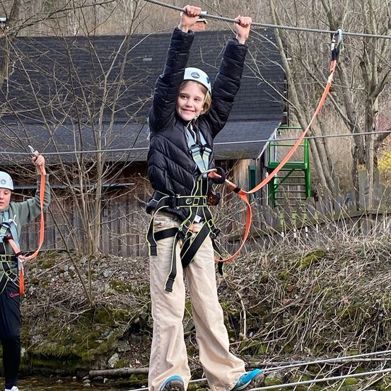 Zwei Personen in Helmen und Gurten halten eine Seilbahn über einem Gewässer. Ein Haus mit einer grünen Treppe befindet sich hinter ihnen.