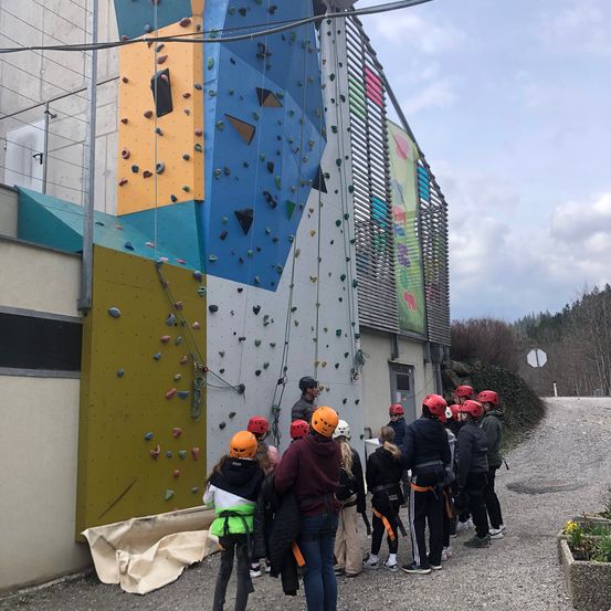 Eine Gruppe von Kindern und Erwachsenen klettert an einer großen Kletterwand, wobei einige Personen bereits klettern und andere auf ihren Zug warten.