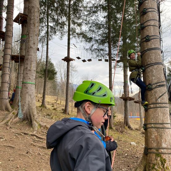 Ein Junge klettert auf einen Baum, während ein anderer Junge zusieht. Der Junge, der den Baum erklimmt, trägt einen grünen Helm und eine Brille. Der zuschauende Junge trägt eine graue Kapuzenjacke und einen grünen Helm.