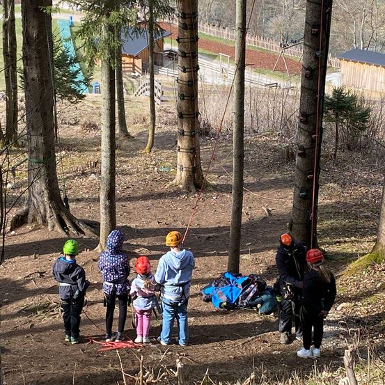 Eine Gruppe von Menschen in einem Wald bereitet sich auf ein Kletterabenteuer vor. Sie tragen Helme und Gurte und stehen neben einem Kletterseil. Einige haben Taschen auf dem Boden.