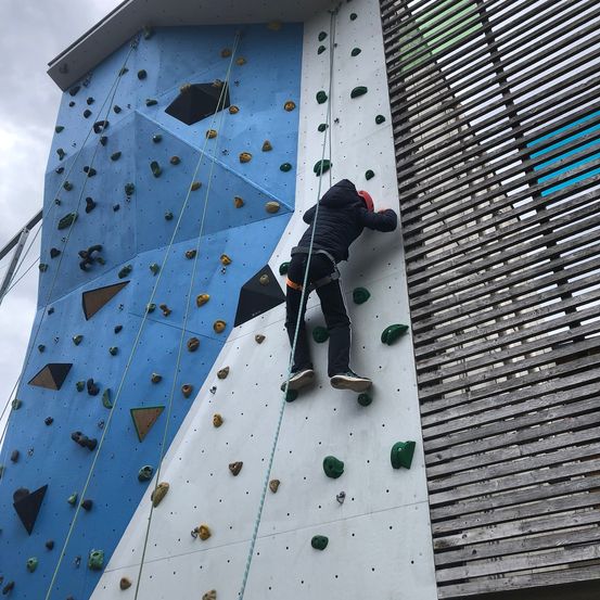 Eine Person klettert an einer blauen und weißen Kletterwand nach oben. Sie trägt eine schwarze Jacke, schwarze Hose und Turnschuhe.