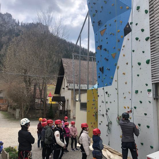 Eine Gruppe von Kindern in roten Helmen und Jacken klettert mit Hilfe eines Lehrers in einer grauen Jacke an einer Kletterwand.