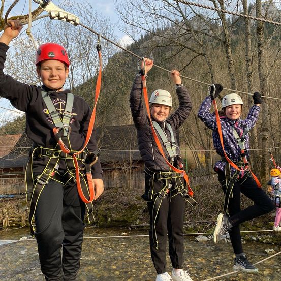 Drei Kinder in Helmen und Gurten, die lächelnd eine Seilbahn über einem Fluss halten, mit Bäumen und einem Gebäude im Hintergrund.