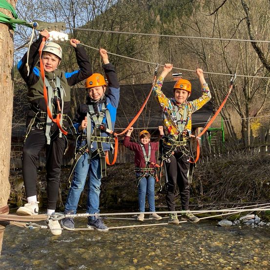 Vier Kinder stehen auf einer Seilbrücke über einem Fluss. Sie tragen Helme und Gurte. Der Junge auf der linken Seite lächelt.