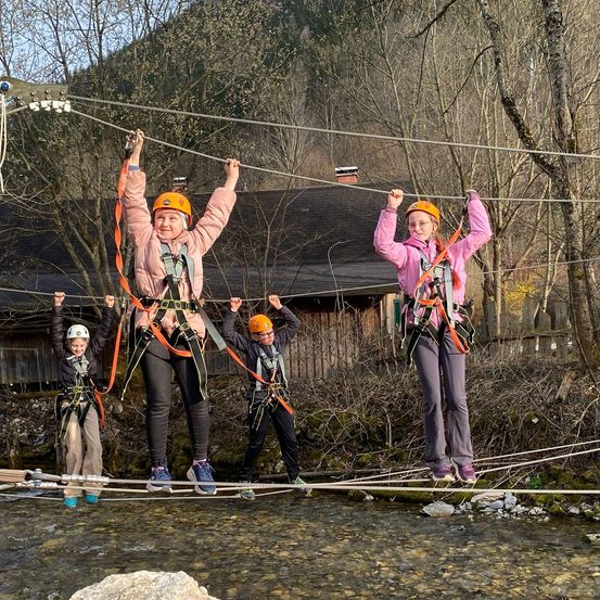 Vier Kinder überqueren eine Hängebrücke über einem Bach. Sie tragen orangefarbene Helme und Sicherheitsgurte. Die Kinder lächeln und scheinen die Aktivität zu genießen.