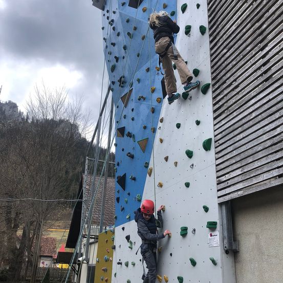 Zwei Personen klettern an einer Kletterwand im Freien. Eine Person trägt einen roten Helm, während die andere einen weißen Helm trägt. Sie sind beide angeschnallt und klettern auf verschiedenen Abschnitten der Wand.