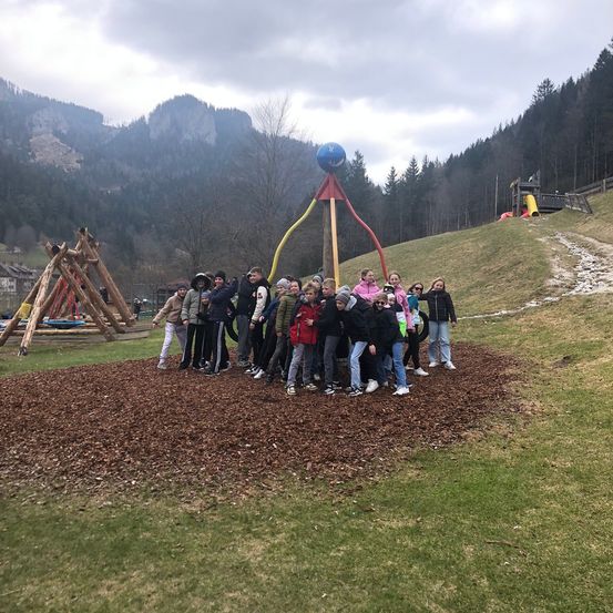 Eine Gruppe von Kindern steht vor einem bunten Metallpfahl in einem Park mit Bergen im Hintergrund.