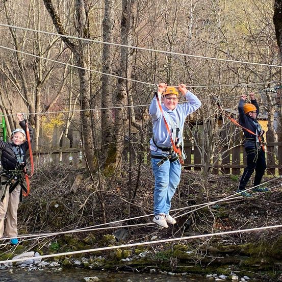 Drei Kinder mit Helmen gehen auf einer Seilbahn in einem Wald.