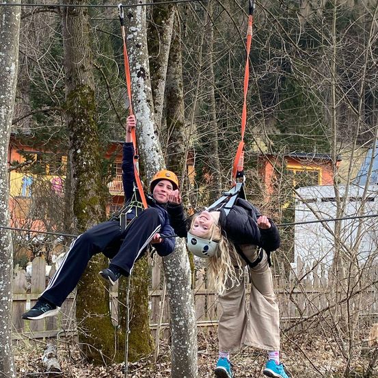 Zwei Personen genießen eine Seilbahnfahrt im Wald, eine trägt einen Helm und die andere einen Rucksack.