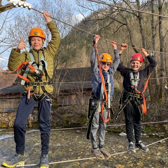 Drei Jungen mit Helmen und Sicherheitsgurten hängen an einer hohen Zipline über einem Fluss.