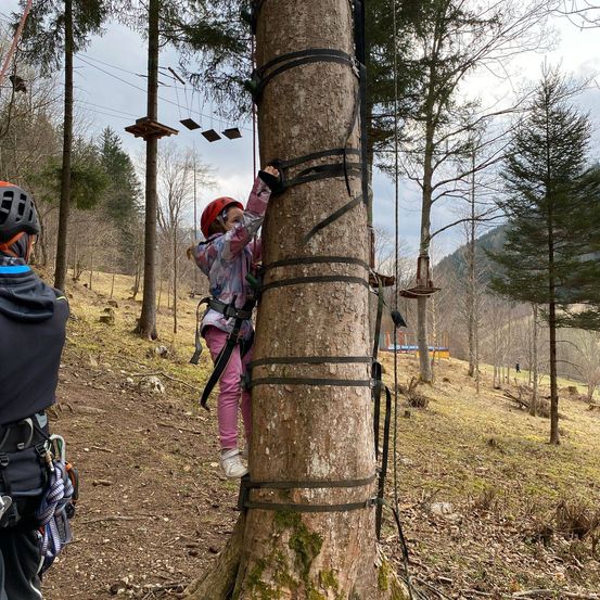 Ein junges Mädchen in einem Helm klettert an einem Baum mit Hilfe von angebrachten Riemen. In der Nähe sieht ein Mann in einem Helm zu.