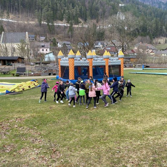 Eine Gruppe von Kindern spielt draußen auf einem Rasenfeld mit einem aufblasbaren Schloss und aufblasbaren Rutschen im Hintergrund.