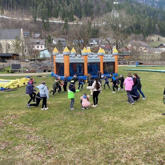 Eine Gruppe von Kindern und Erwachsenen spielt auf einem Rasenfeld mit Bergen im Hintergrund um einen blauen und orangefarbenen Hüpfburg.