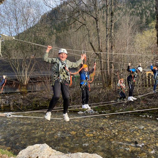 Mehrere Kinder und Jugendliche überqueren eine Hängebrücke über einem Fluss. Sie tragen Sicherheitsausrüstungen.