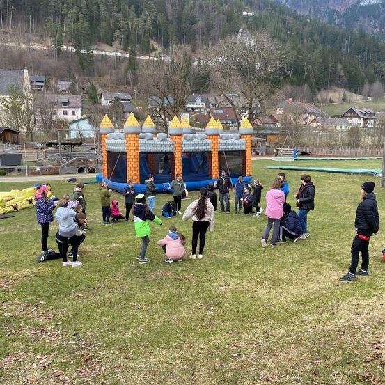 Eine Gruppe von Menschen auf einem Feld mit einem großen aufblasbaren Schloss dahinter. Im Hintergrund befinden sich Häuser mit einem Bergrücken.