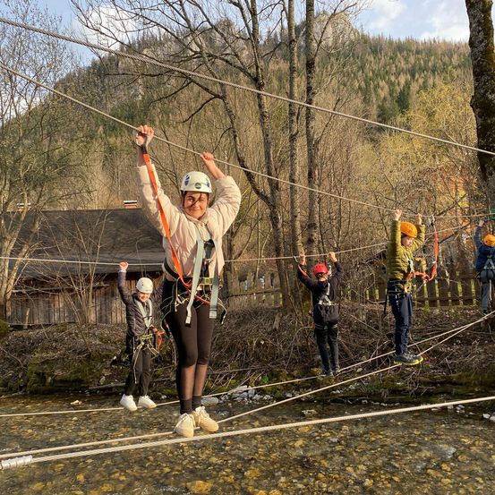 Menschen in Sicherheitsausrüstung und Helmen überqueren ein Seil über einem Fluss.