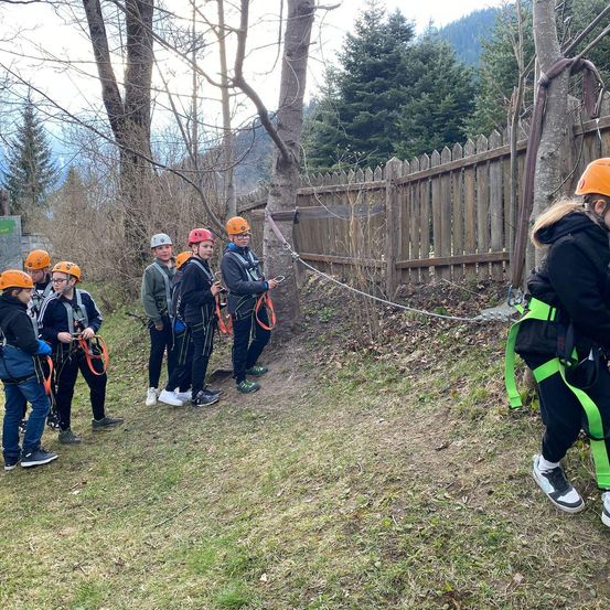 Schüler in Helmen stehen in einem Grasbereich in der Nähe eines Zauns und warten wahrscheinlich auf eine Kletteraktivität.
