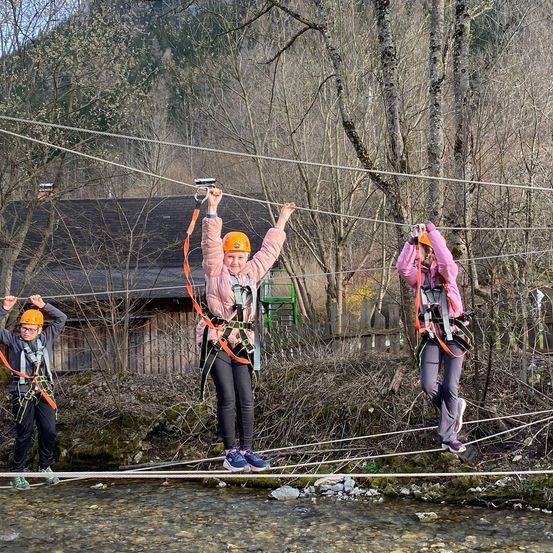 Drei Personen gehen auf einer aufgehängten Brücke über einen Fluss. Alle tragen Helme und Gurte. Die Brücke besteht aus Seilen und wird von Bäumen getragen.