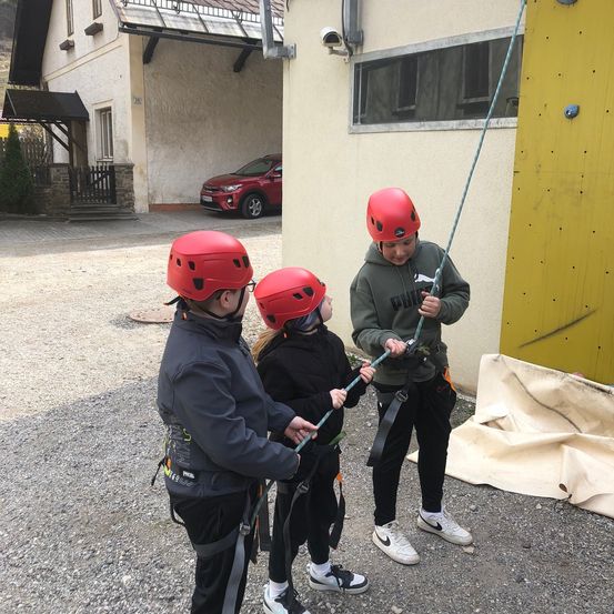 Drei Kinder stehen vor einem Gebäude mit einer Garage, in der ein Auto steht. Sie tragen rote Helme und Gurte.