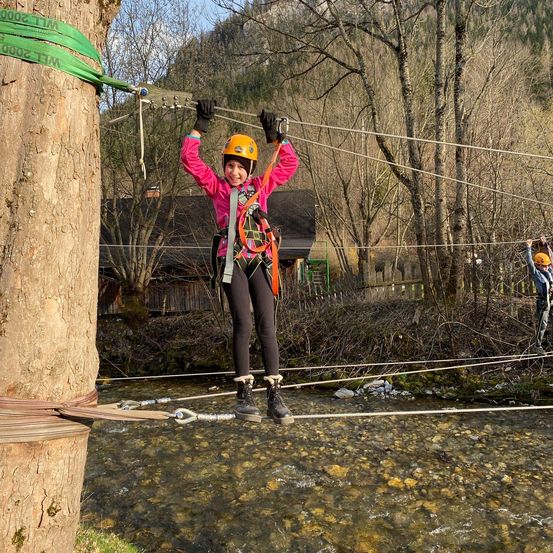 Ein junges Mädchen in gelbem Helm und Sicherheitsgurt geht über ein Seil über einem Fluss.