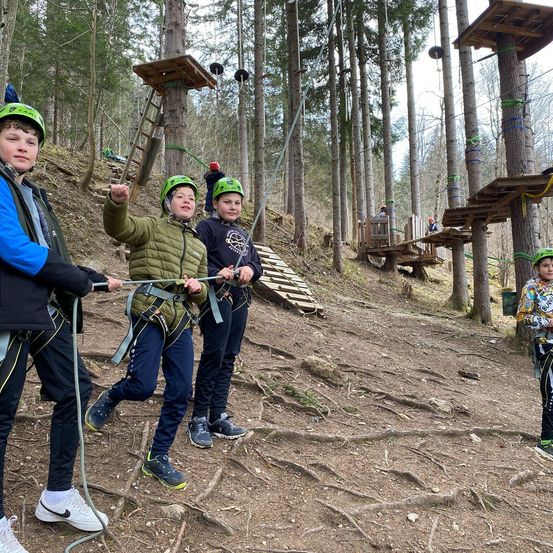 Mehrere Kinder bereiten sich auf ein Abenteuer im Wald vor. Sie tragen Helme und halten Seile. Sie stehen auf einem Hang mit Baumwurzeln. In der Ferne befinden sich Holzkonstruktionen mit Leitern.