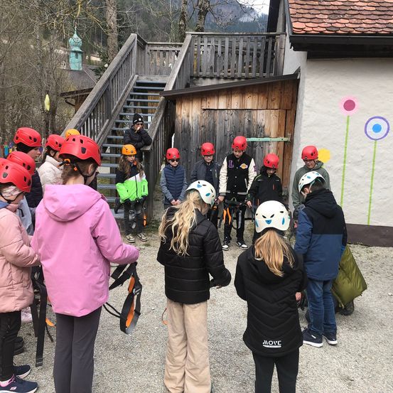 Eine Gruppe von Kindern mit roten Helmen steht draußen, einige tragen Jacken und Turnschuhe. Sie machen wahrscheinlich eine kurze Pause vor dem Start einer Aktivität.