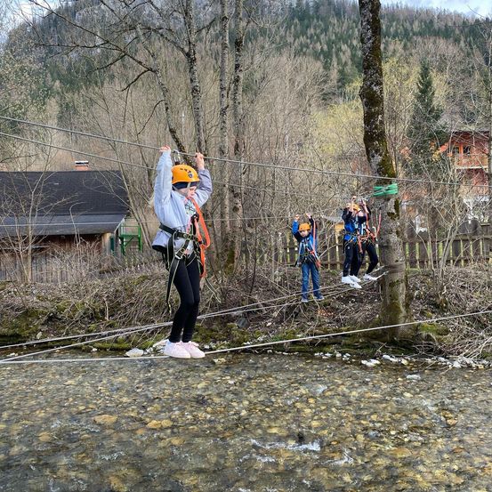 Mehrere Personen fahren auf einer Seilbahn über einen Fluss, tragen Helme und Sicherheitsausrüstung. Dahinter steht ein Holzhaus und ein Gebäude mit Balkon.