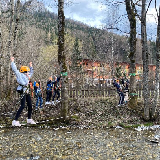 Eine Gruppe von Menschen in Helmen und Gurten überquert eine Hängebrücke über einem kleinen Bach.