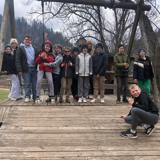 Eine Gruppe von Kindern und Jugendlichen posiert auf einer Holzplattform für ein Foto, einige halten einen Stock. Sie sind in Winterkleidung gekleidet.